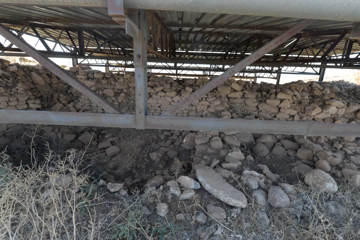 A low timber shelter spans a stone excavation area in the open archaeological site.