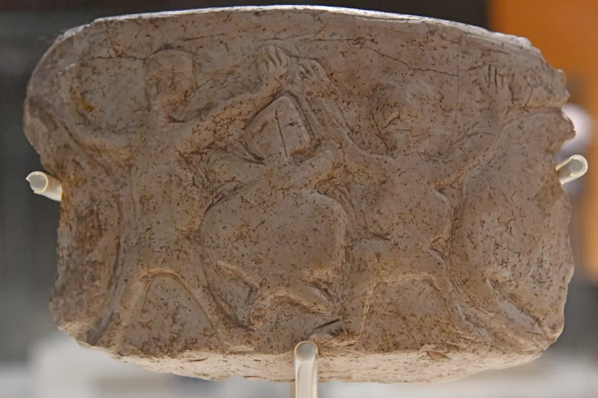 An oblong pale stone vessel fragment with carved figures across its surface, mounted on white supports in a museum case.